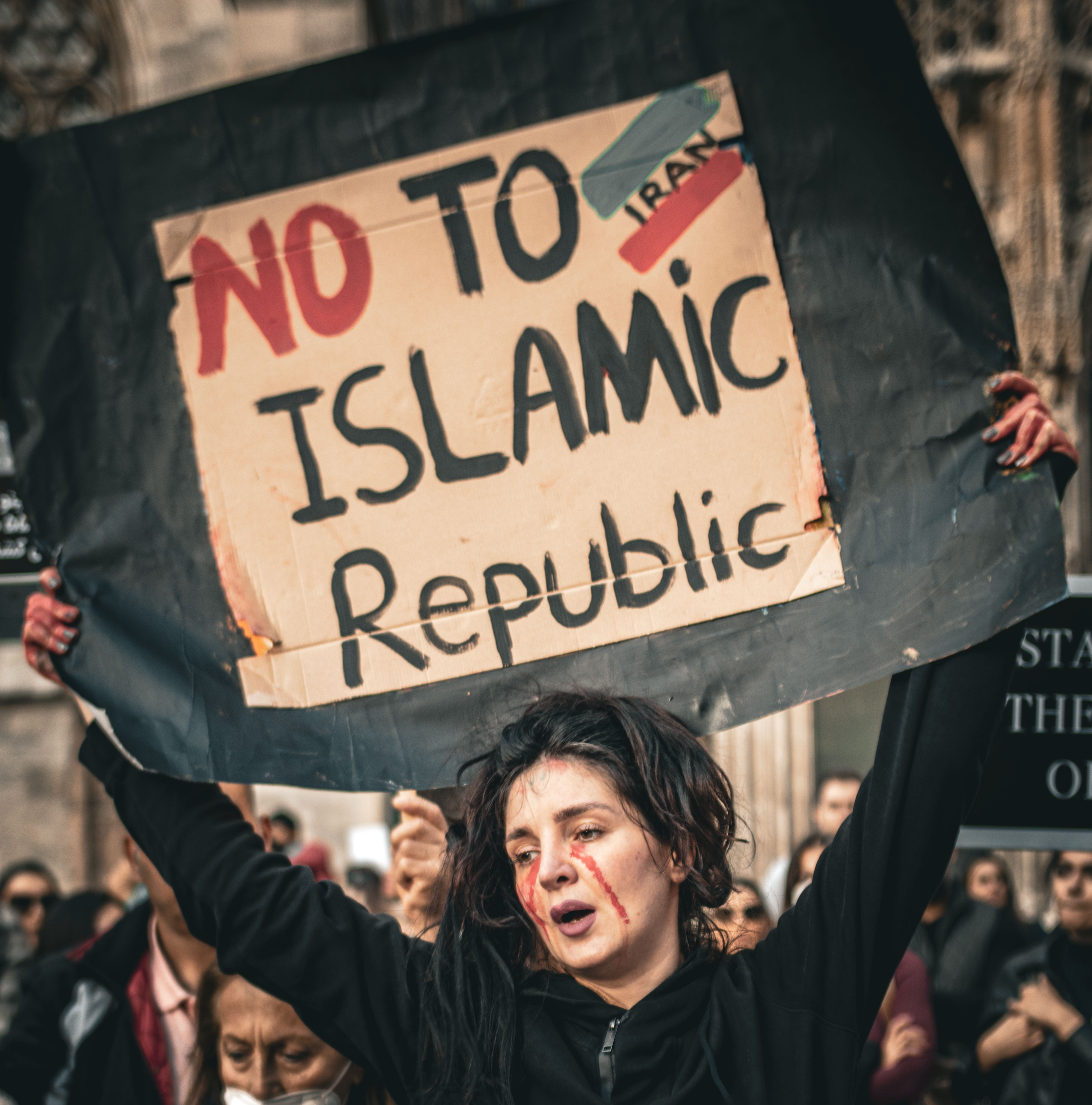 Photograph by Babak Dalivand, in Austria, Vienna, 2022. An Iranian woman is pictured protesting in front of St. Stephen's Cathedral in Vienna. She has dark hair and a black sweatshirt and is wearing red face paint in the appearance of bloody tears; her mouth is slightly agape. She holds a cardboard sign as she looks downwards and toward the right. A crowd can be seen behind her.