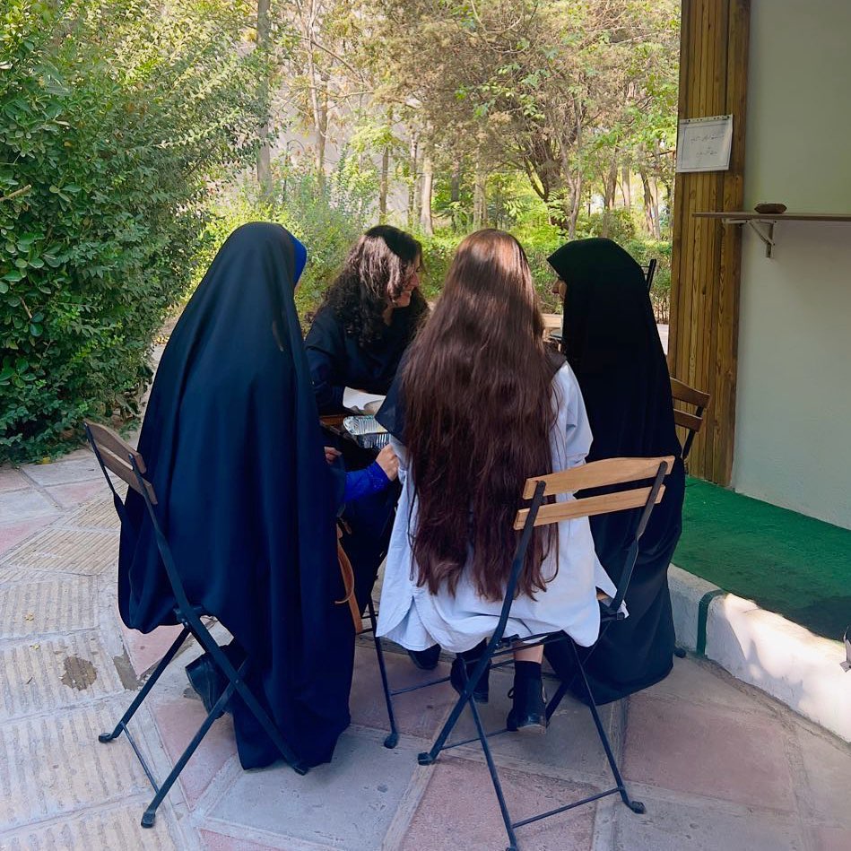 Photograph from 2022 Mahsa Amini Uprising; four women chat pleasantly at a table in Iran. Two are wearing conservative black chadors; two are without hijab.